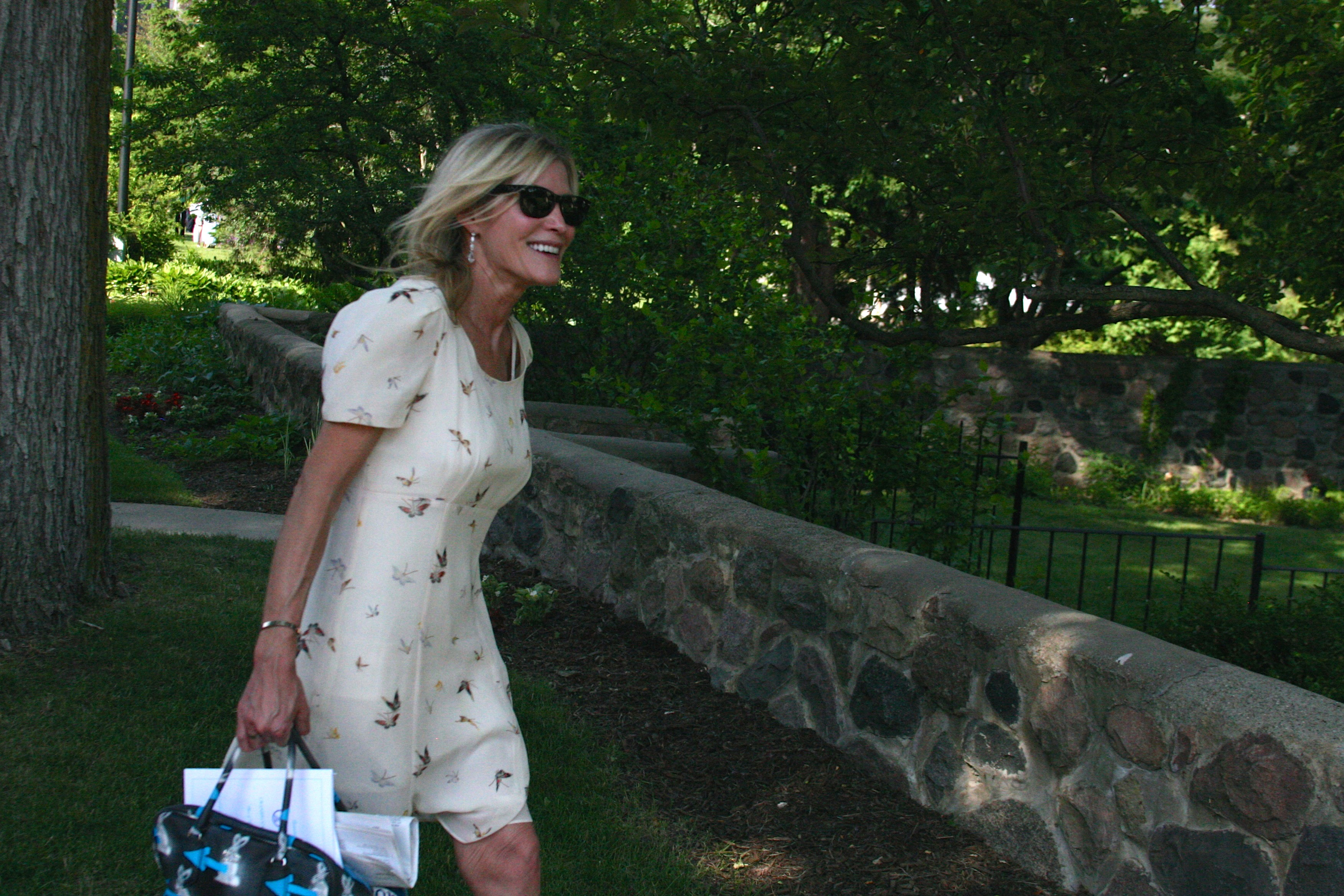 Kogler set against a backdrop of trees, smiling and holding a purse and wearing sunglasses