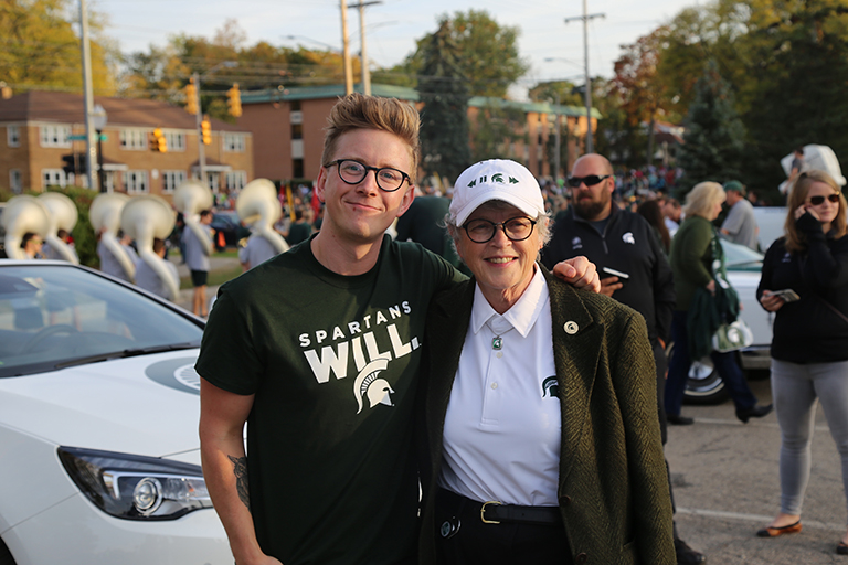 ComArtSci alumnus Tyler Oakley stands in front of his parade car with his arm around MSU President Simon.